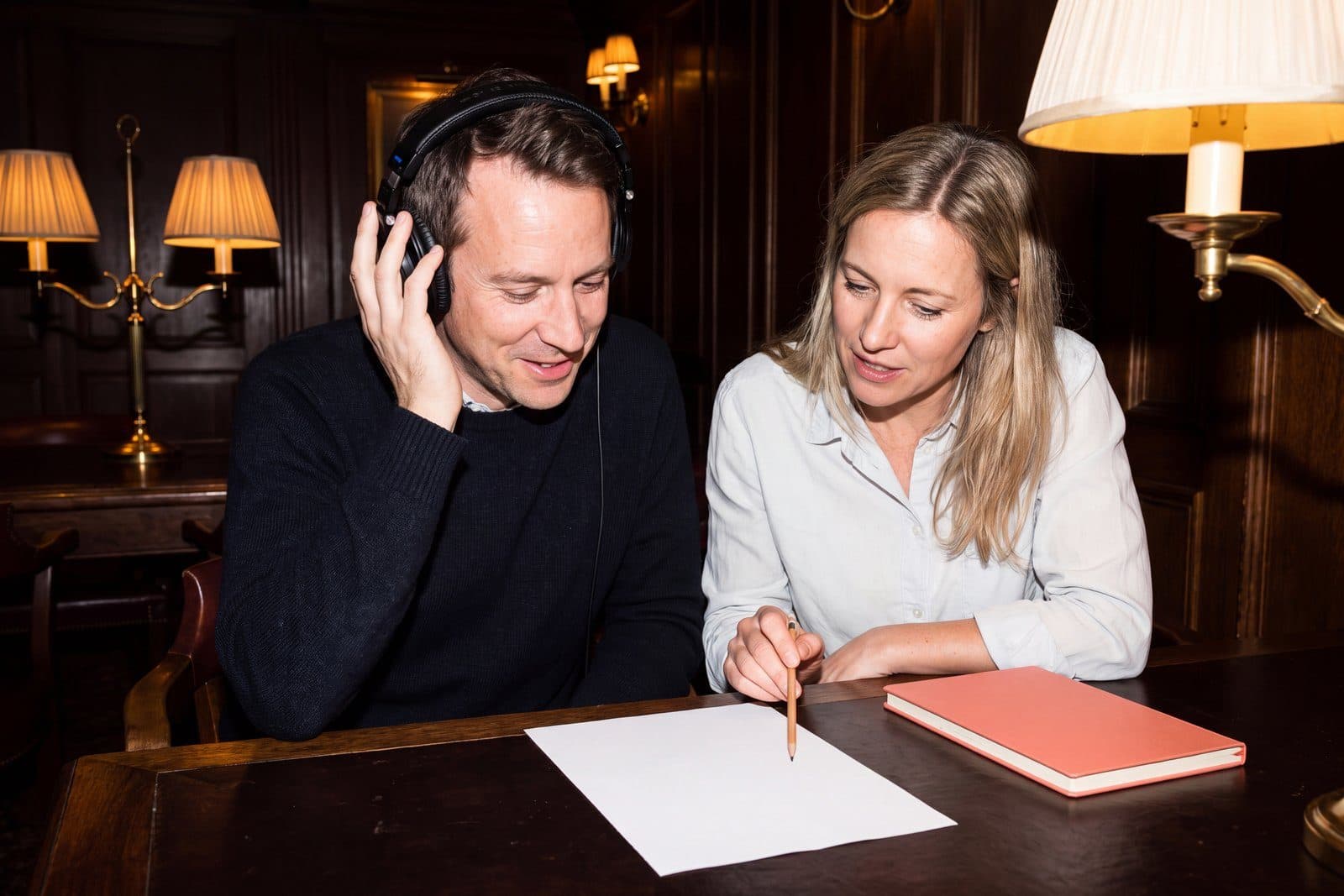 Two people at a dark wood desk in a club room, one listening on headphones while the other points a pencil at a blank sheet beside a coral notebook